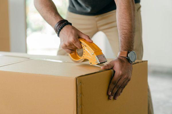 Person sealing a cardboard box with packing tape preparing for a move or placing items into self storage during a life event transition